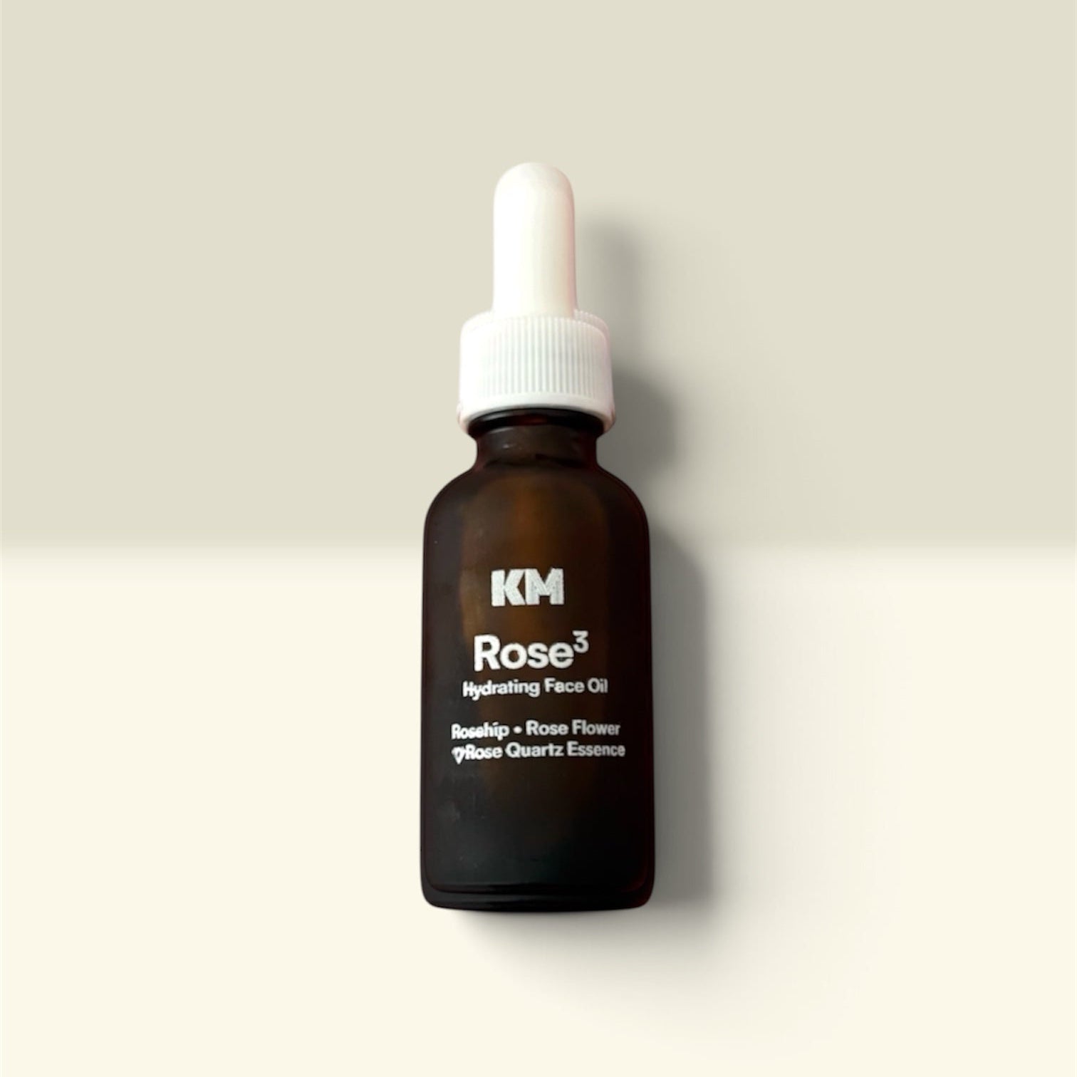 KM Rose Hydrating Face Oil in an amber glass dropper bottle with a white cap, centered against a warm beige background.