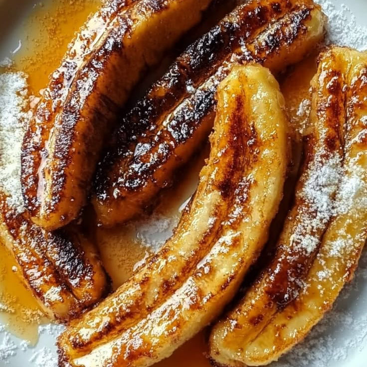 Caramelized banana slices on a white plate, drizzled with syrup and dusted with powdered sugar.